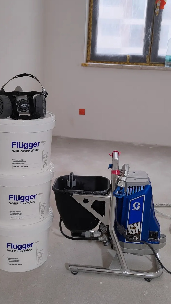 Sprzęt do natryskowego nakładania farby Graco GX FF obok trzech wiader z gruntem Flügger Wall Primer White i maski ochronnej w pomieszczeniu z oknem zabezpieczonym folią.