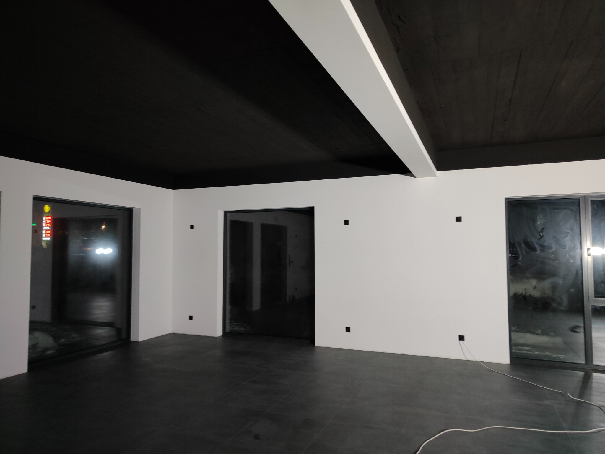 Wnętrze po remoncie z pomalowanym na czarno sufitem z widoczną fakturą drewna, białymi ścianami i ciemną podłogą. Widoczne okna i drzwi z czarnymi ramami.