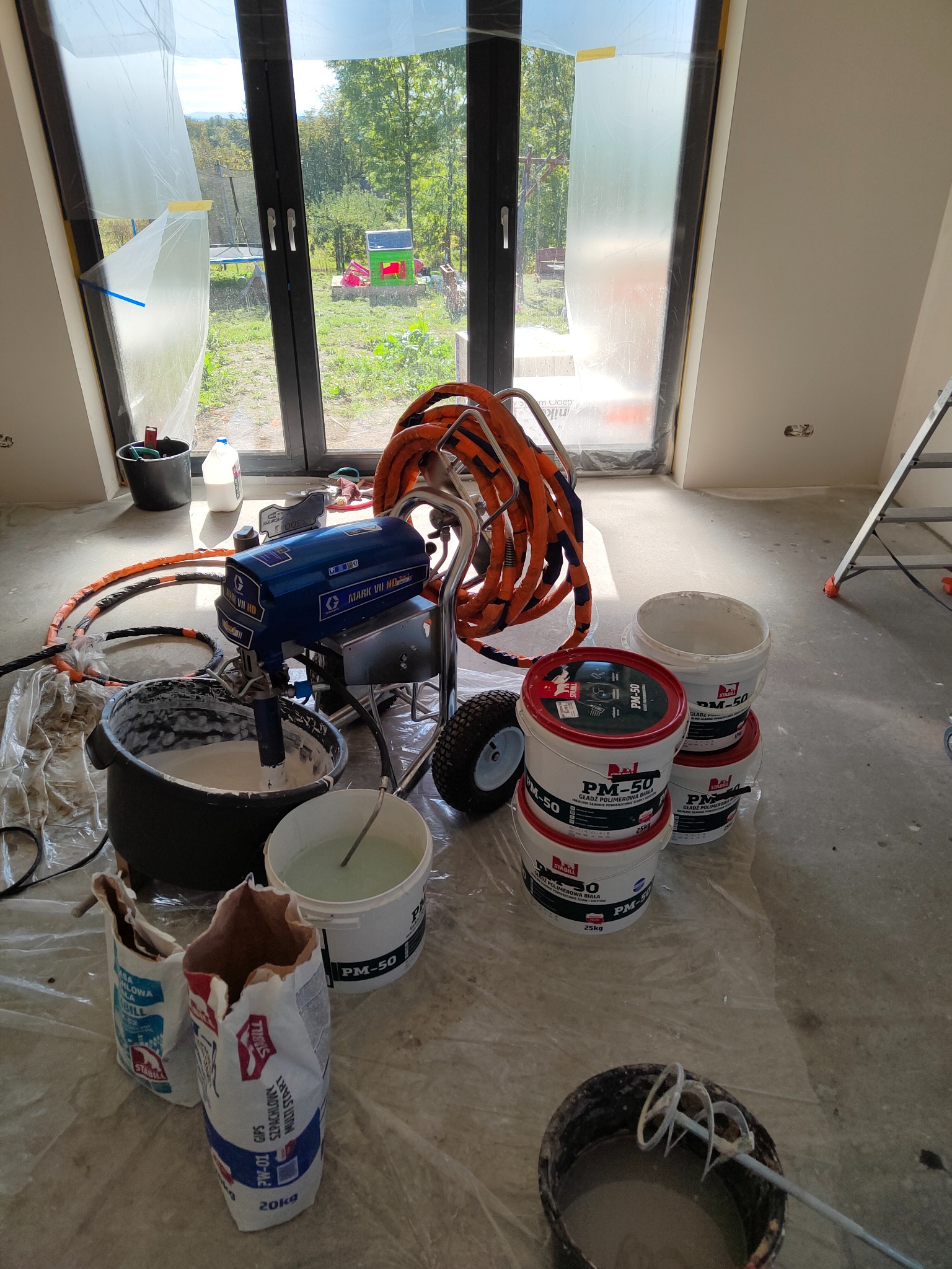 Przygotowanie do prac wykończeniowych: agregat do natrysku gładzi, wiadra z gotową masą polimerową PM-50 oraz worki z gipsem szpachlowym na podłodze zabezpieczonej folią malarską, widoczne okno...
