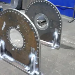 Dwie metalowe konstrukcje z otworami i śladami po spawaniu, ustawione na betonowej podłodze w warsztacie, z widocznymi elementami wyposażenia w tle.