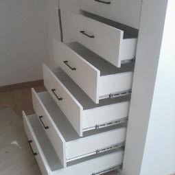 Biała szafa z wysuniętymi szufladami, czarne uchwyty, widoczne prowadnice, minimalistyczny design, jasne wnętrze szuflad.