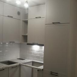 Jasna kuchnia z białymi, kafelkowymi ścianami i szarymi blatami. Minimalistyczne szafki z ciemnymi uchwytami. Dwa kryształowe źródła światła.