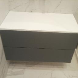 Szara szafka łazienkowa z białym blatem, wisząca na marmurowej ścianie. Minimalistyczny design, nowoczesne wykończenie, widoczne dwie szuflady.
