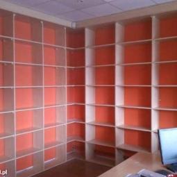 Pusta, narożna szafa wnękowa z jasnego drewna i pomarańczowym tłem, widoczna z perspektywy wnętrza pomieszczenia, oświetlona światłem z góry.