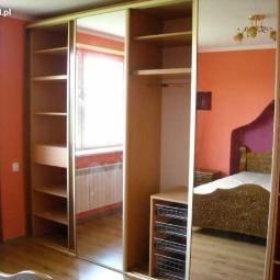 Beżowa szafa przesuwna z lustrami w pokoju o pomarańczowych ścianach. Widoczne półki i szuflady wewnątrz szafy. Elementy wystroju wnętrza odbijają się w lustrach.