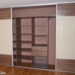 Wnętrze szafy wnękowej z przesuwnymi drzwiami w odcieniach brązu i beżu, widoczne półki, szuflady i kosz na ubrania. Jasna podłoga z paneli.