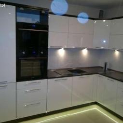 Błyszcząca, biała kuchnia na wymiar z czarnym piekarnikiem i oświetleniem LED pod szafkami oraz na podłodze. Minimalistyczny design, nowoczesne wykończenie.