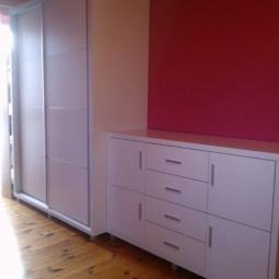 Biała szafa przesuwna i komoda z szufladami na drewnianej podłodze, na tle ściany w kolorze kremowym i bordowym. Ujęcie z boku, widoczne detale wykończenia.