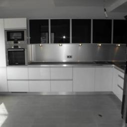 Nowoczesna kuchnia z białymi szafkami dolnymi i czarnymi górnymi. Metalowy panel nad blatem, wbudowane sprzęty AGD. Minimalistyczny design, oświetlenie punktowe.