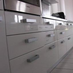 Biała, lakierowana zabudowa kuchenna z szufladami i srebrnymi uchwytami. U góry wbudowany piekarnik marki Bosch. Minimalistyczny styl, jasne wnętrze.
