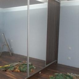 Szafa przesuwna z lustrzanymi drzwiami, widoczna drabina i materiały budowlane w pokoju, brązowe panele na podłodze, szare ściany, minimalistyczny design.