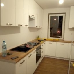 Jasna kuchnia z białymi, lakierowanymi szafkami i drewnianym blatem. Na blacie poziomica i butelka wody. Widoczne okno z nocnym widokiem.