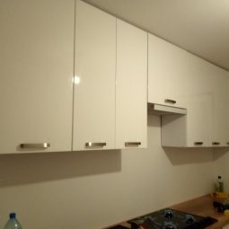 Białe, lakierowane szafki kuchenne wiszące, z minimalistycznymi uchwytami, oświetlone punktowo. Widok na fragment blatu i płyty grzewczej. Nowoczesny design.
