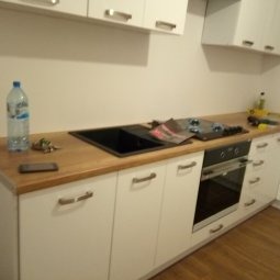 Biała, minimalistyczna kuchnia z drewnianym blatem, czarnym zlewem i srebrnymi uchwytami. Na blacie butelka wody i płyta grzewcza. Ujęcie z niskiego kąta.