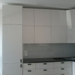 Biała, nowoczesna zabudowa kuchenna z połyskującymi frontami, czarnym blatem i szarą ścianą nad blatem. Minimalistyczne uchwyty podkreślają prostotę designu.