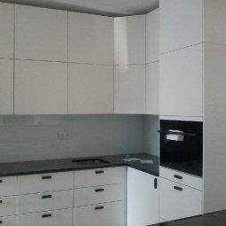 Biała, minimalistyczna zabudowa kuchenna z czarnym blatem i dyskretnymi uchwytami, wkomponowana w jasne wnętrze. Widoczny piekarnik w zabudowie.