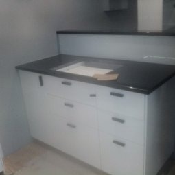 Biała szafka kuchenna z czarnym blatem i minimalistycznymi uchwytami, widoczna w rogu pomieszczenia, z kartonem na blacie. Nowoczesny design.