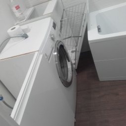 Biała pralka ładowana od przodu w małej łazience z brązową podłogą. Nad pralką papier toaletowy i płyn do prania. Obok kosz na pranie i wanna.