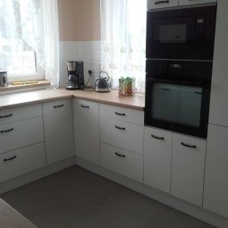 Jasna kuchnia z białymi frontami i drewnianym blatem, czarne uchwyty, piekarnik w zabudowie, ekspres do kawy na blacie, widok na okno z firanką.