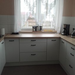 Jasna kuchnia z białymi szafkami i drewnianym blatem pod oknem, widok na zewnątrz. Na blacie akcesoria kuchenne, w tle firanka w oknie.