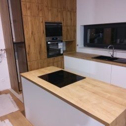 Nowoczesna kuchnia z wyspą: białe fronty szafek dolnych i drewnopodobne górne. Czarna płyta indukcyjna na blacie wyspy, okno nad zlewem. Minimalistyczny design.