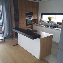 Nowoczesna kuchnia z wyspą: drewno i biel, okno z tulipanami, czarny blat wyspy na metalowej konstrukcji. Minimalistyczny regał z książkami w tle.