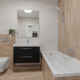 Terra Estate - biuro nieruchomości - Nowoczesna łazienka z wanną, umywalką z szafką i toaletą w jasnych płytkach imitujących drewno. Minimalistyczny design, lustro i roślina doniczkowa.
