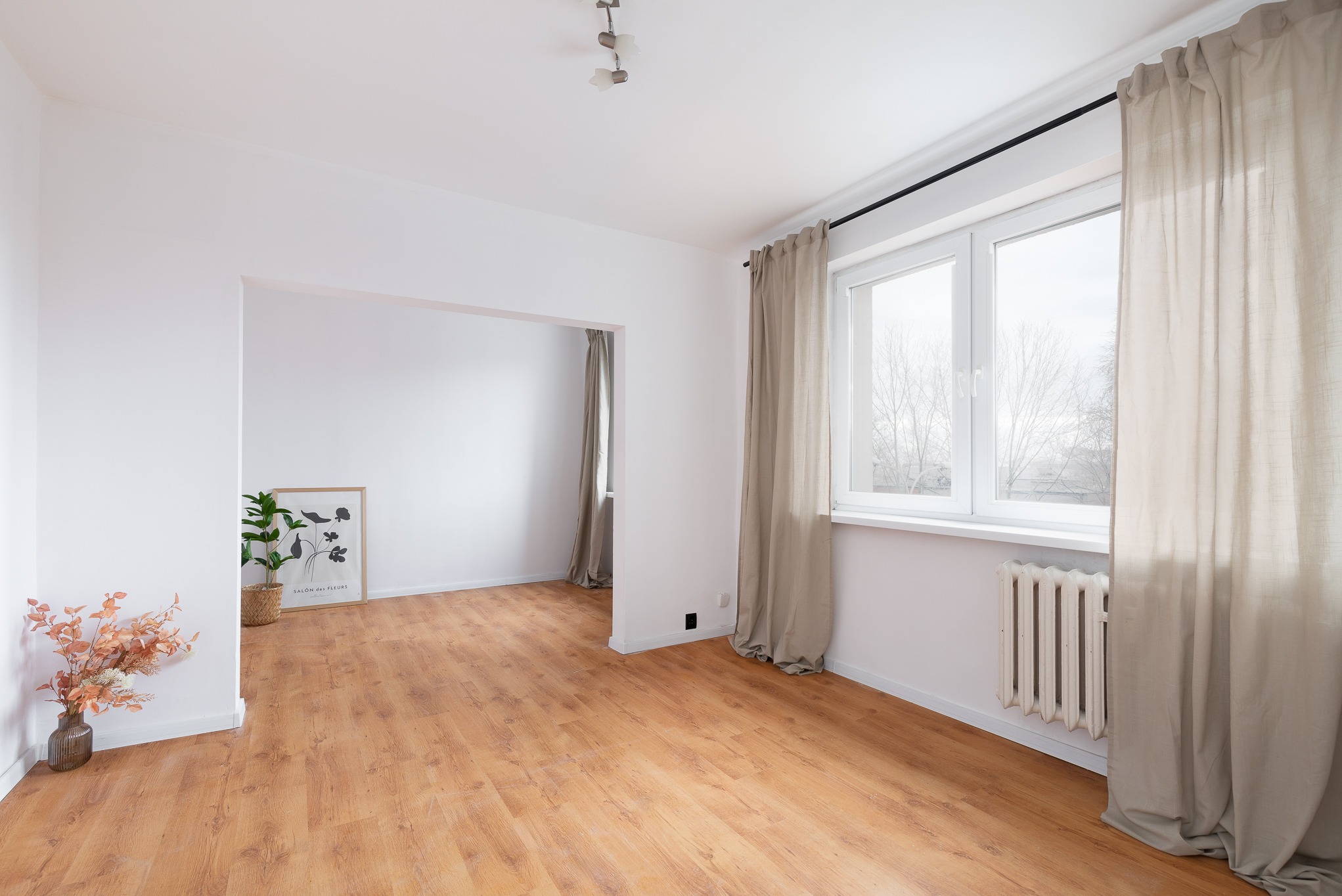 Jasne, puste wnętrze z drewnianą podłogą, białymi ścianami, oknem z beżowymi zasłonami i dekoracjami roślinnymi. Widok na sąsiednie pomieszczenie.