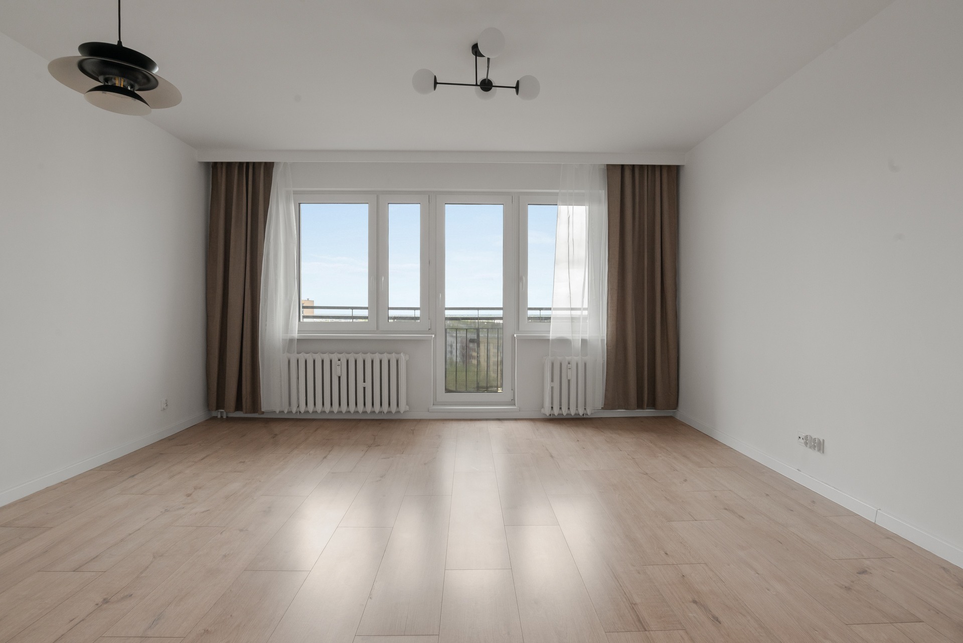 Jasne, puste wnętrze z drewnianą podłogą i dużym oknem balkonowym z widokiem na miasto. Minimalistyczne lampy sufitowe i brązowe zasłony dodają elegancji.
