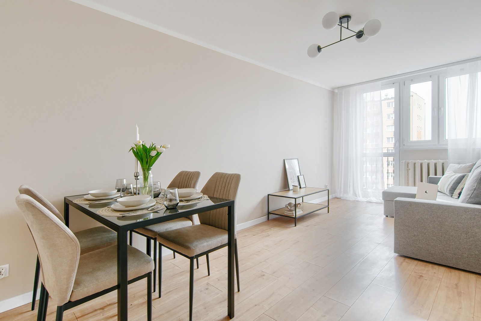 Jasne wnętrze mieszkania z jadalnią: stół z nakryciem, krzesła z tapicerką, sofa i stolik kawowy. Minimalistyczny design, drewniana podłoga.