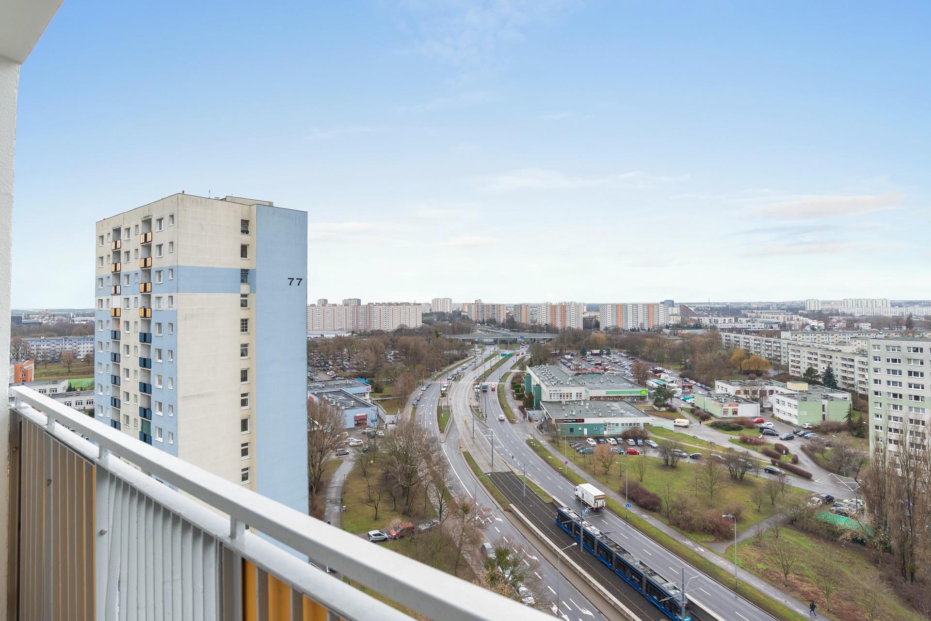 Widok z balkonu na miasto z blokami, ulicą i tramwajem. Blok z numerem 77 po lewej stronie. Jasne niebo.