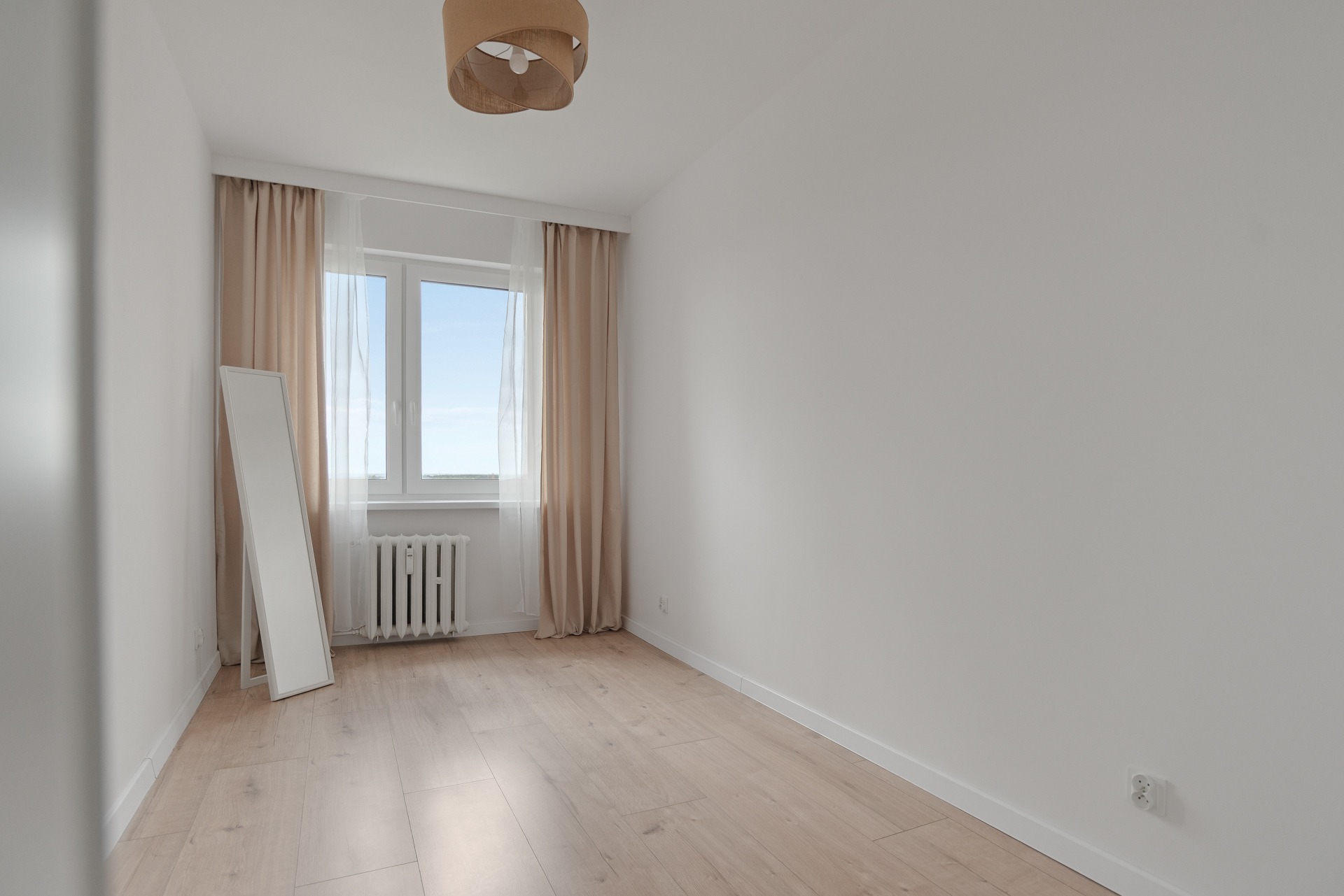 Jasne, puste wnętrze pokoju z drewnianą podłogą, grzejnikiem, oknem z zasłonami i dużym lustrem opartym o ścianę. Neutralna kolorystyka ścian i wyposażenia.