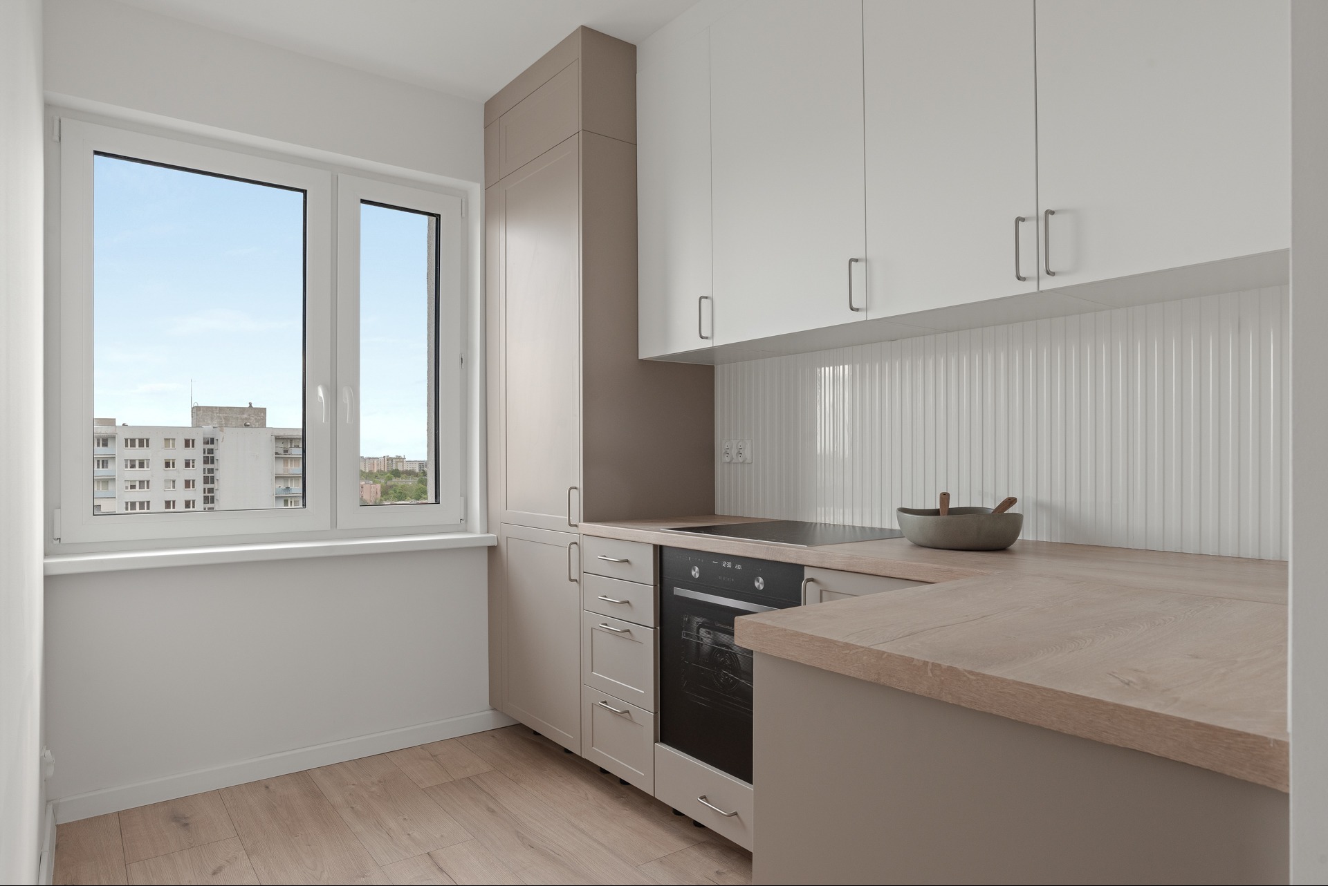 Jasna kuchnia z drewnianym blatem i widokiem na miasto z okna. Minimalistyczny design, neutralne kolory, nowoczesne wyposażenie.