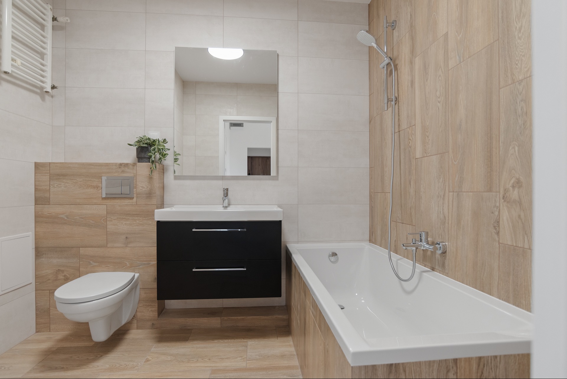 Nowoczesna łazienka z wanną, umywalką z szafką i toaletą w jasnych płytkach imitujących drewno. Minimalistyczny design, lustro i roślina doniczkowa.
