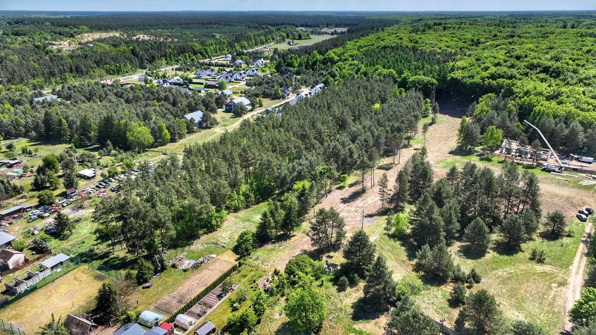 Panoramiczny widok z lotu ptaka na osiedle domów jednorodzinnych otoczone lasem, z placem budowy w prawym dolnym rogu. Widoczne maszyny budowlane i fundamenty nowego domu.