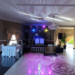 Piotr Wałowski - Maverick Guy Music - Sala bankietowa z przygotowanym stanowiskiem DJ-a: biały podświetlany stół, sprzęt nagłośnieniowy, oświetlenie sceniczne i dekoracyjne, kryształowy żyrandol, stolik z zastawą i kwiatowe dekoracje...