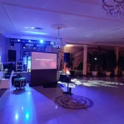 Sala bankietowa przygotowana na imprezę, widoczne oświetlenie sceniczne, projektor wyświetlający tekst 'Gdzie te prywatki nie zapomniane', nagłośnienie, stoły z białymi obrusami i dekoracyjne...
