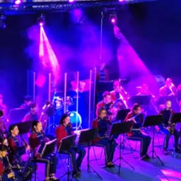 Orkiestra dęta w czerwono-niebieskich mundurach gra na scenie oświetlonej fioletowymi reflektorami, z widocznymi instrumentami dętymi i perkusyjnymi.