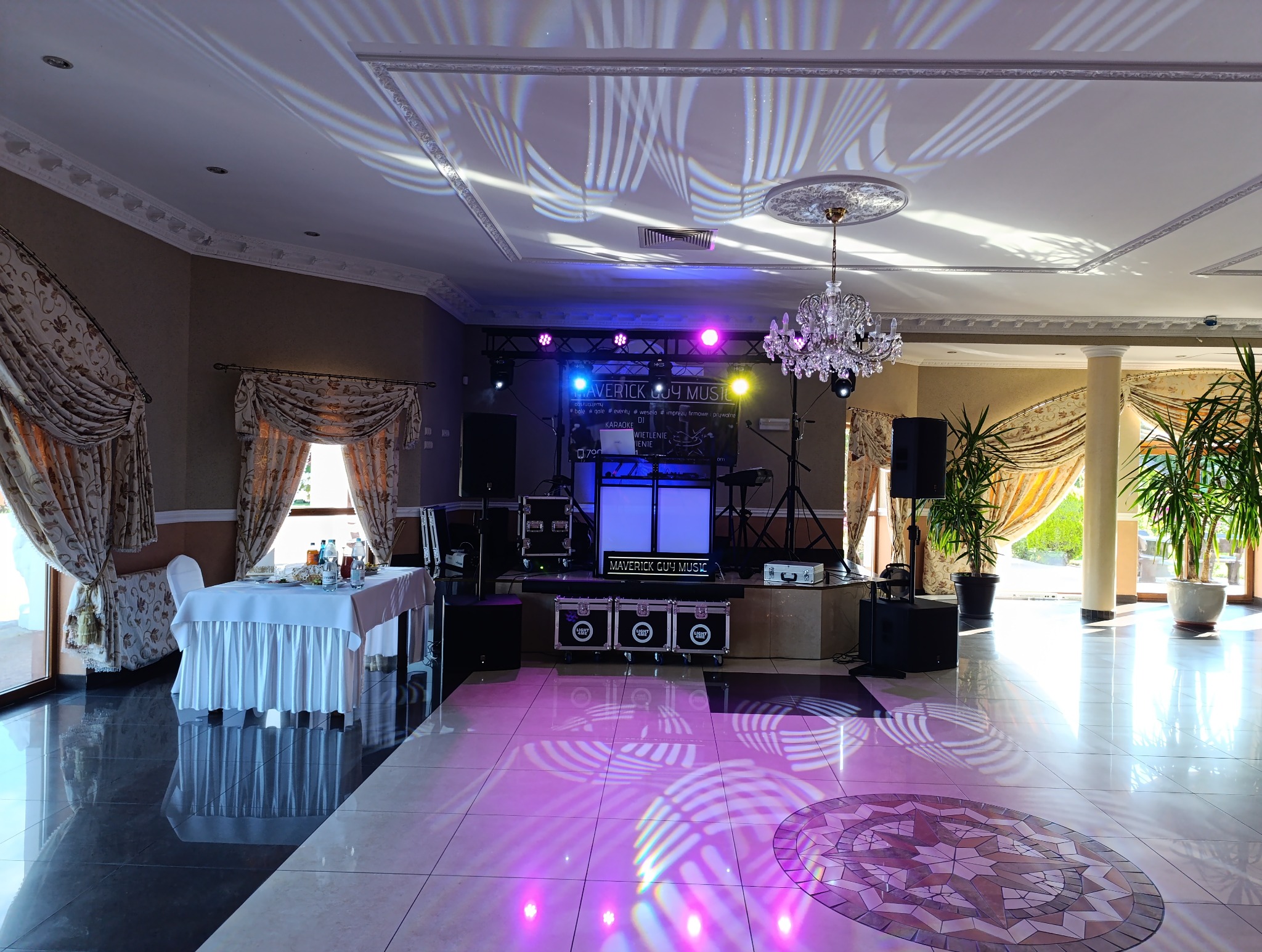 Sala bankietowa z przygotowanym stanowiskiem DJ-a: biały podświetlany stół, sprzęt nagłośnieniowy, oświetlenie sceniczne i dekoracyjne, kryształowy żyrandol, stolik z zastawą i kwiatowe dekoracje...