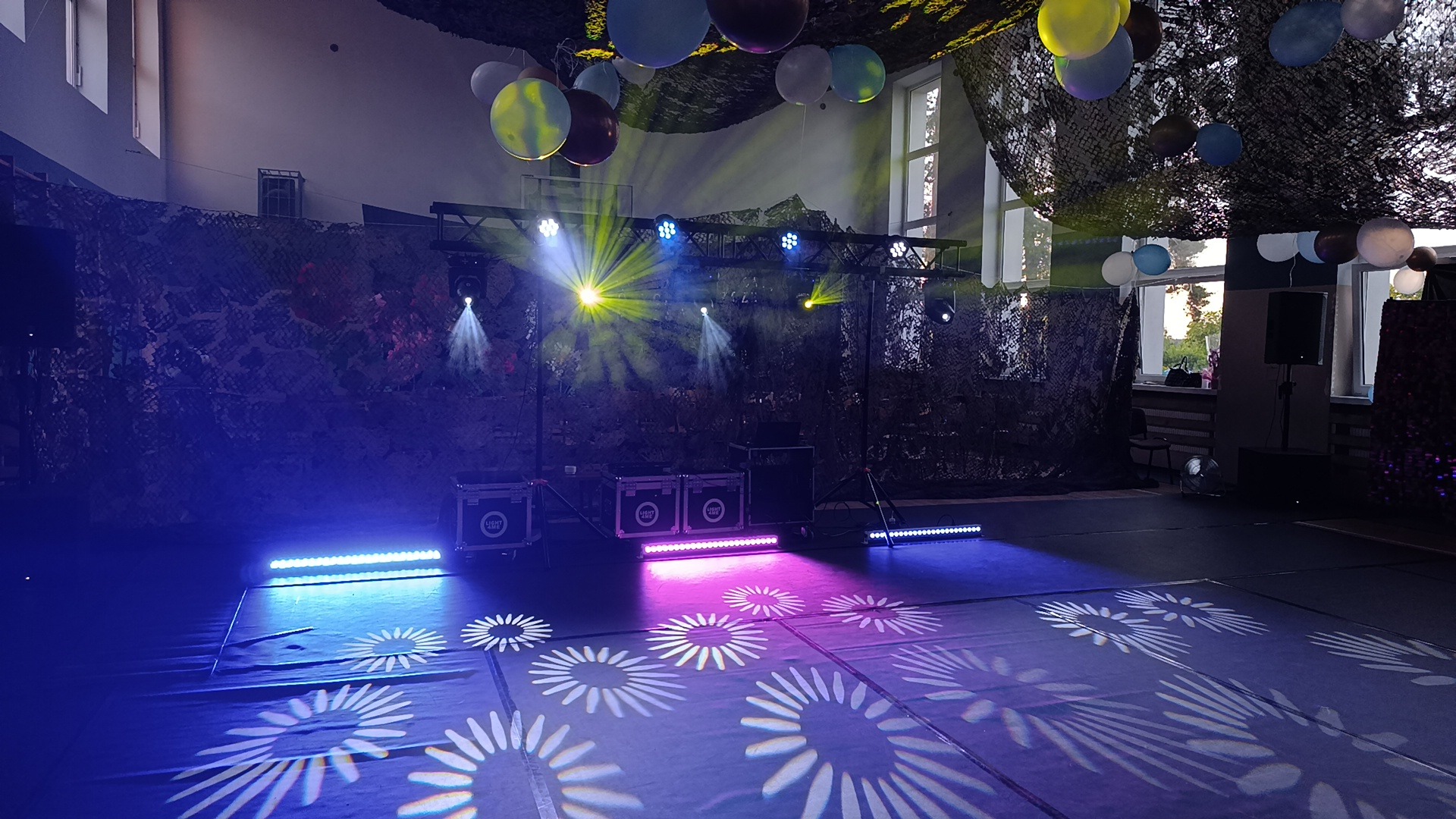 Sala udekorowana na imprezę z profesjonalnym oświetleniem: reflektory, belki LED, dekoracje z siatki maskującej i balonów.