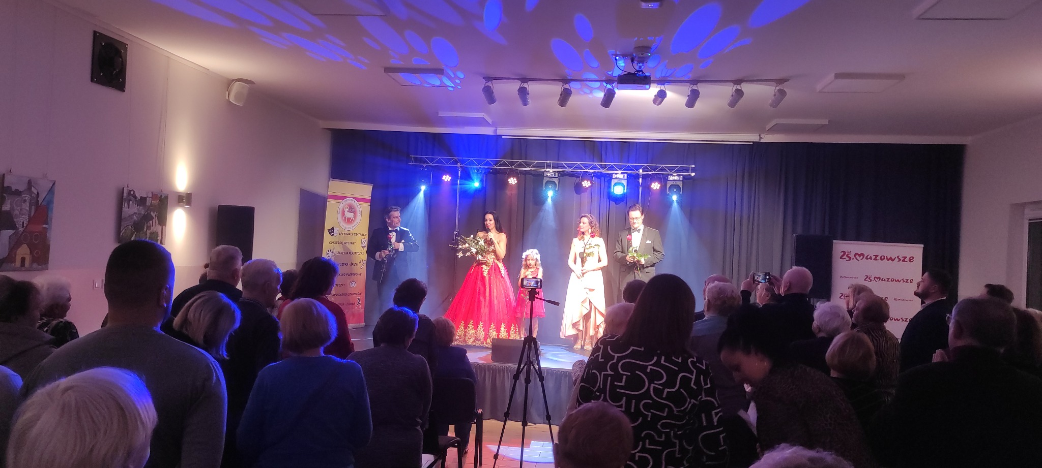 Występ na scenie podczas uroczystości, oświetlenie punktowe, dwoje prezenterów w strojach galowych, kobieta w czerwonej sukni, publiczność obserwująca wydarzenie, widoczny baner reklamowy w tle.
