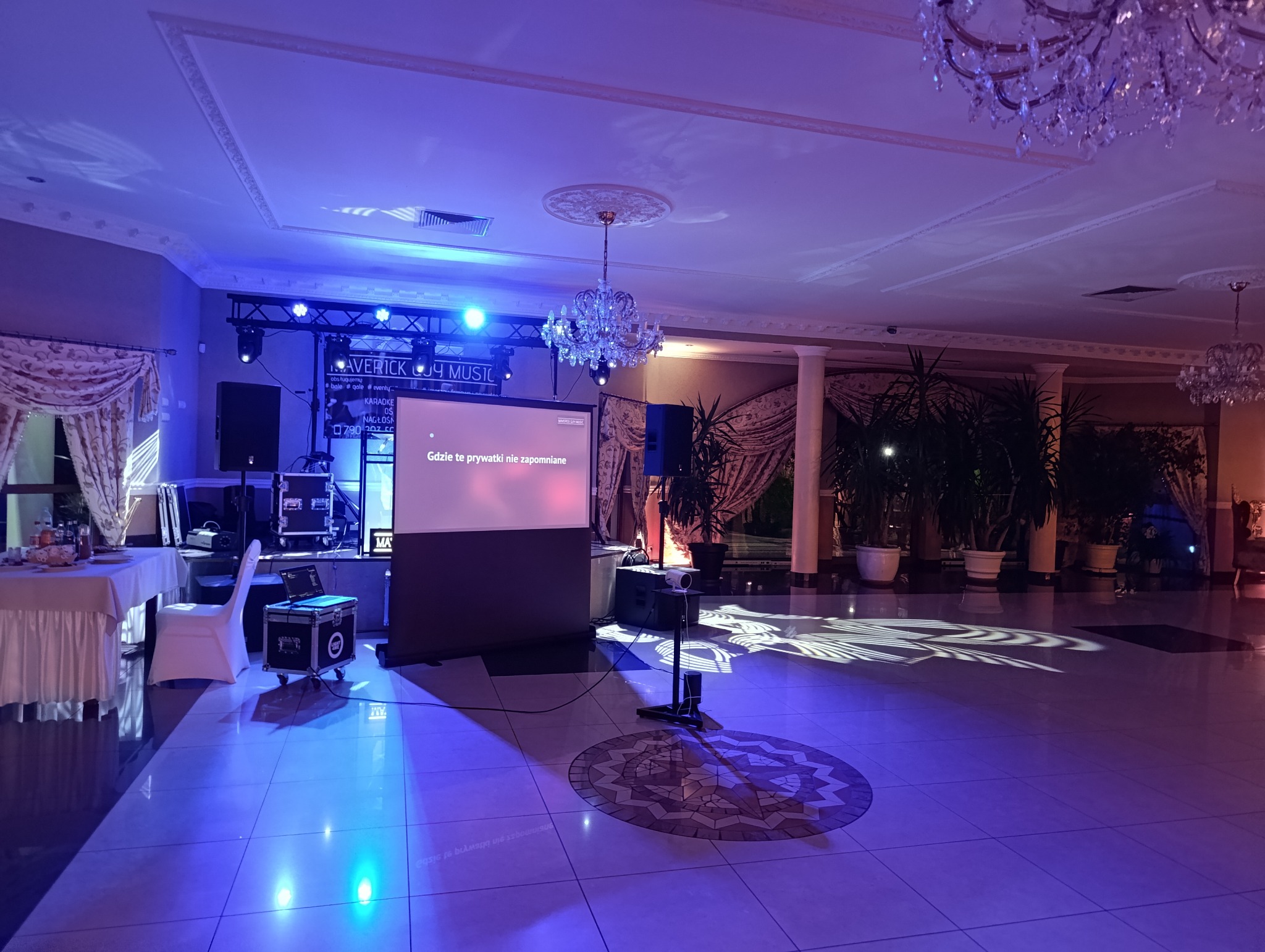 Sala bankietowa przygotowana na imprezę, widoczne oświetlenie sceniczne, projektor wyświetlający tekst 'Gdzie te prywatki nie zapomniane', nagłośnienie, stoły z białymi obrusami i dekoracyjne...