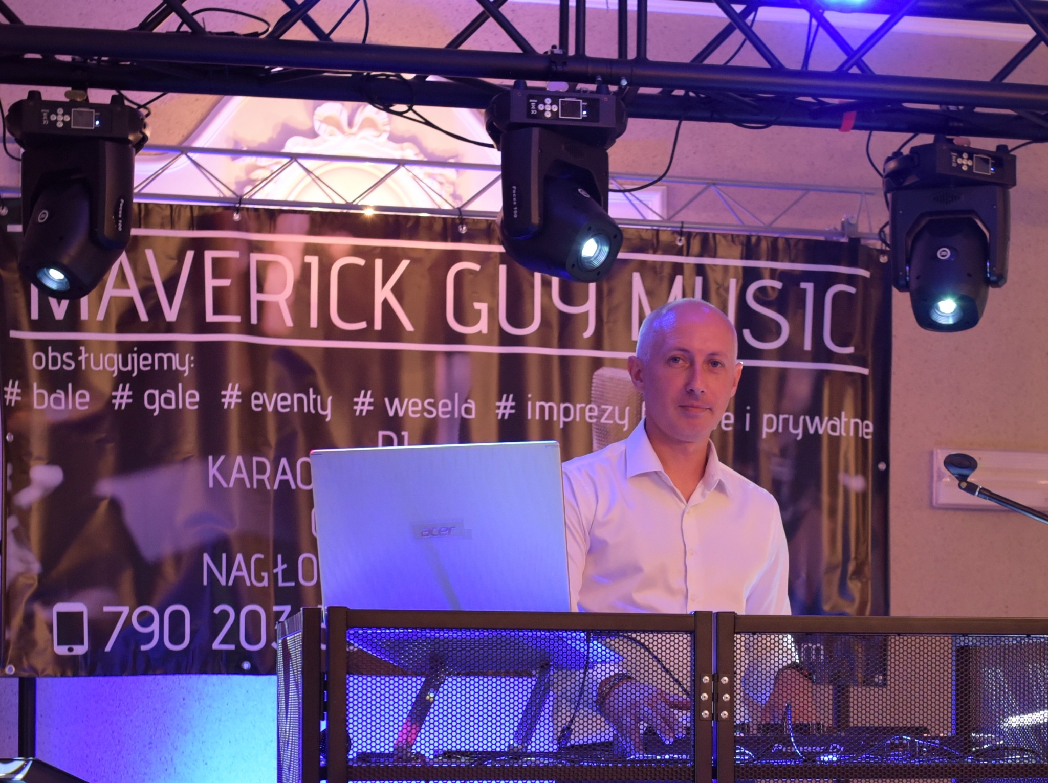 DJ za konsoletą z laptopem Acer, oświetlenie sceniczne i baner reklamowy z numerem telefonu. Obsługa imprez, bale, gale, wesela.