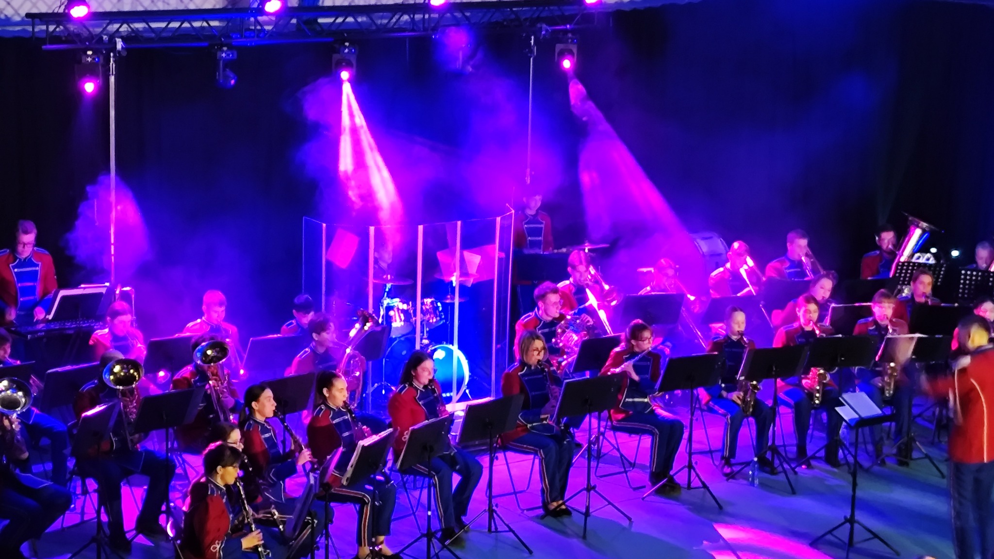 Orkiestra dęta w czerwono-niebieskich mundurach gra na scenie oświetlonej fioletowymi reflektorami, z widocznymi instrumentami dętymi i perkusyjnymi.