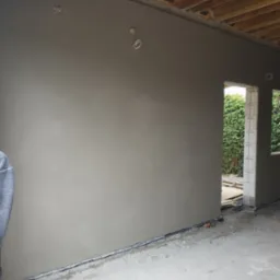 Świeżo nałożony, surowy tynk maszynowy na ścianie wewnętrznej, widoczne puszki elektryczne pod gniazdka, surowy strop i widok na zieleń przez otwór okienny.