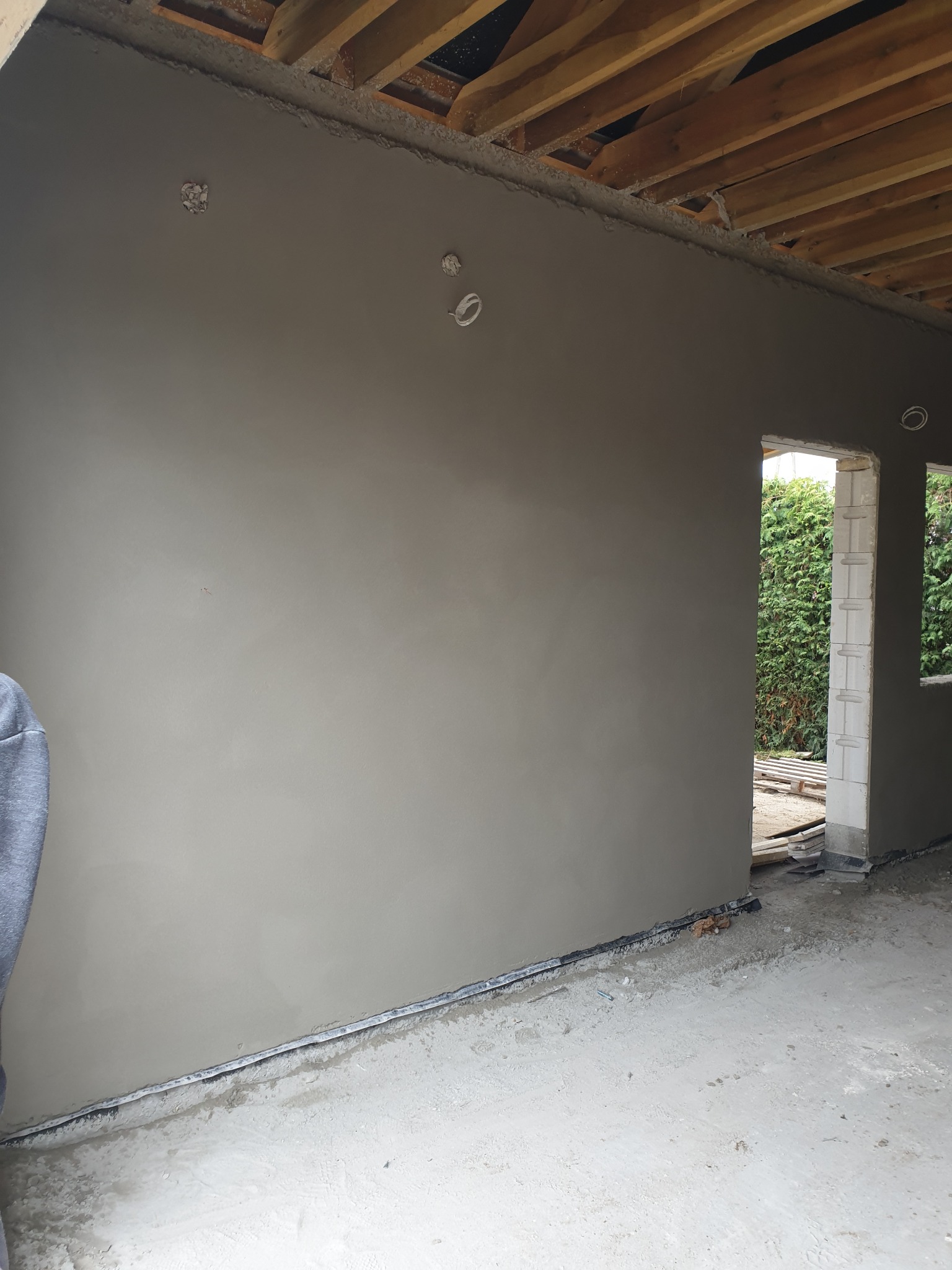 Świeżo nałożony, surowy tynk maszynowy na ścianie wewnętrznej, widoczne puszki elektryczne pod gniazdka, surowy strop i widok na zieleń przez otwór okienny.