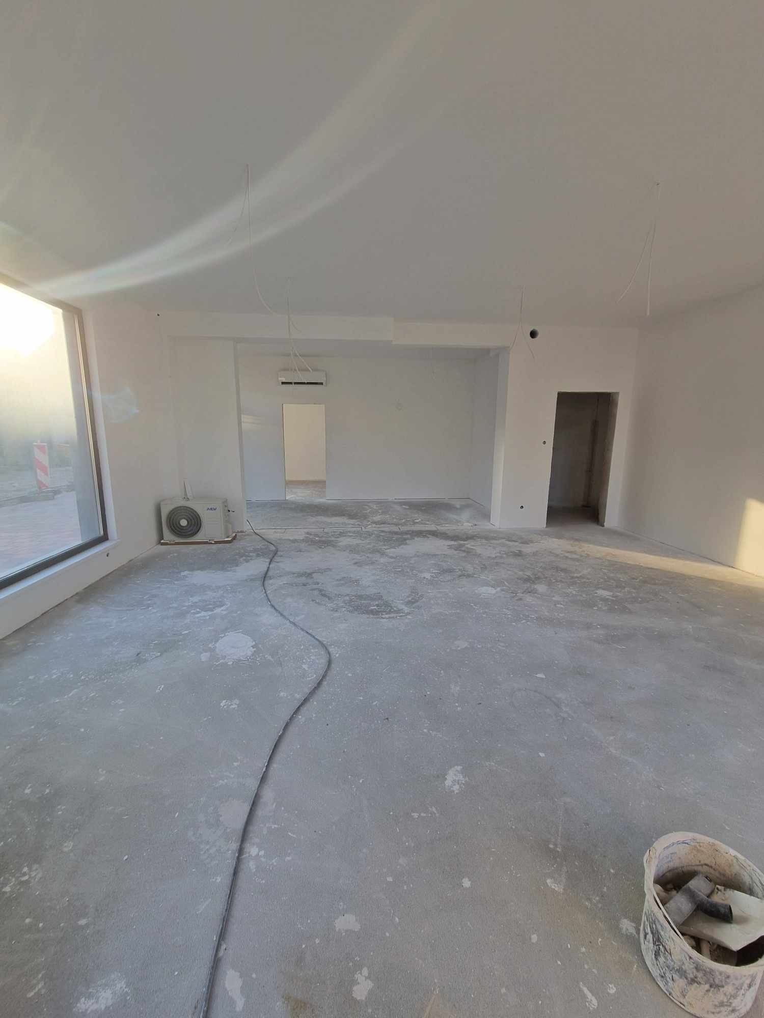 Wnętrze w trakcie prac wykończeniowych: świeżo wykończone ściany, widoczne przewody, klimatyzator przy oknie i wiadro z narzędziami na podłodze.