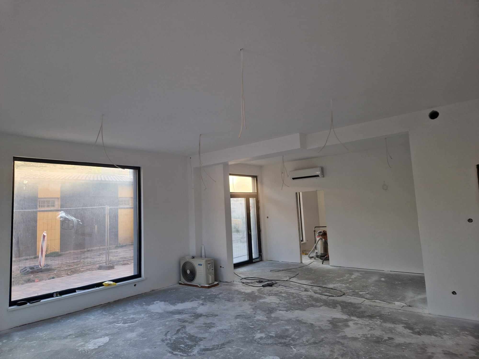 Wnętrze w trakcie prac wykończeniowych: gładkie, białe ściany, surowy beton na podłodze, widoczne przewody wystające z sufitu, okno z widokiem na zewnątrz i klimatyzator w rogu pomieszczenia.