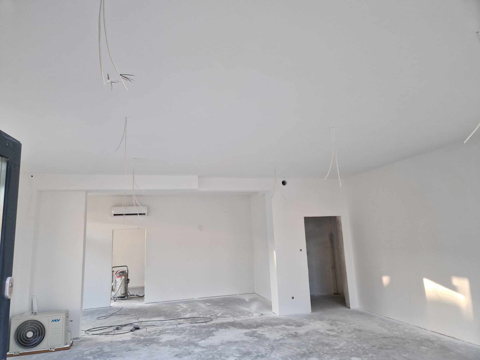 Przygotowane do prac wykończeniowych wnętrze: białe ściany, surowa podłoga, wystające kable i klimatyzator, widoczny odkurzacz przemysłowy w tle.