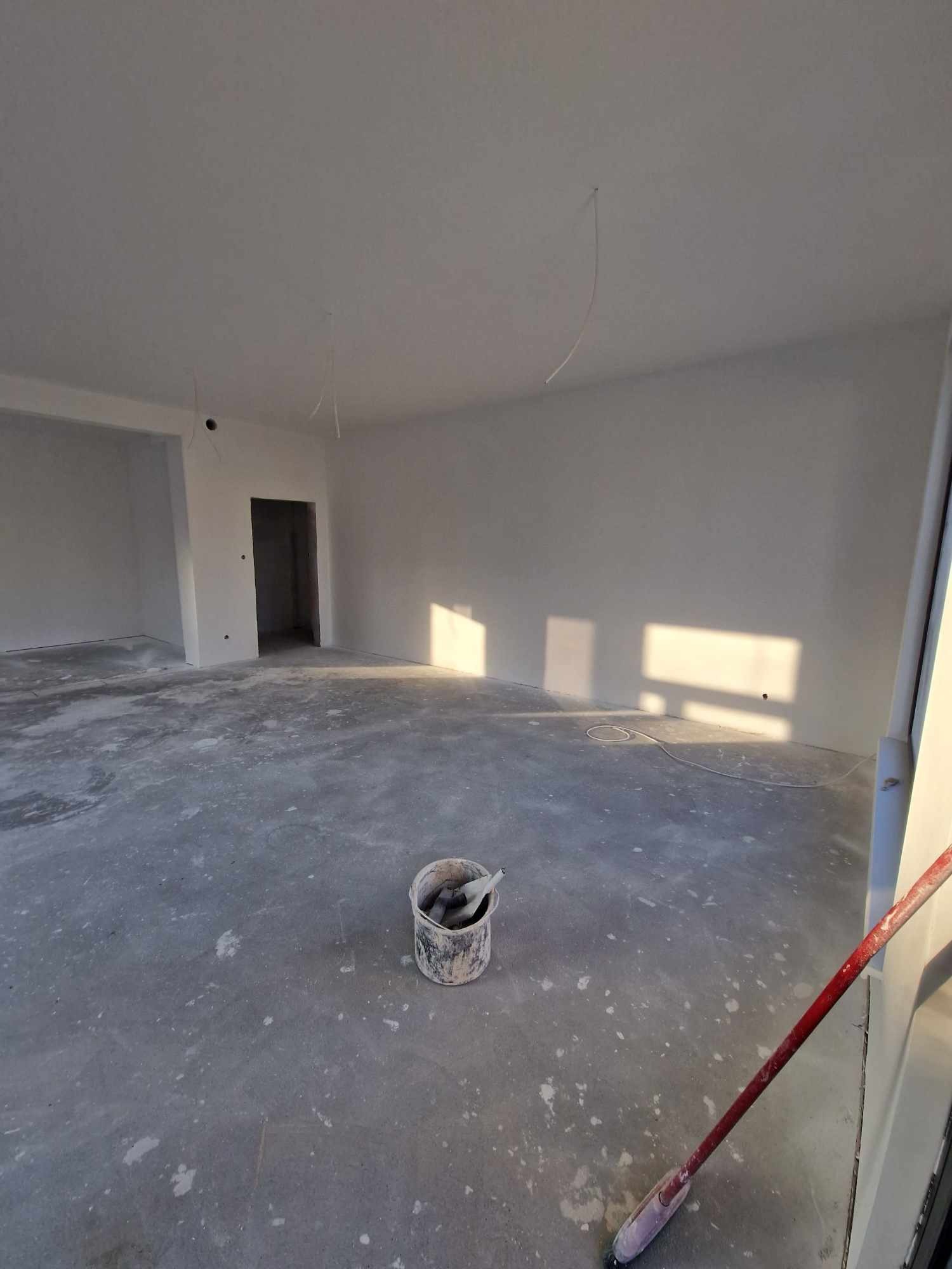 Wnętrze w trakcie prac wykończeniowych: surowe ściany w jasnym odcieniu, z widocznymi śladami po kablach, betonowa posadzka z plamami farby, wiadro z narzędziami malarskimi i wałek malarski w rogu...